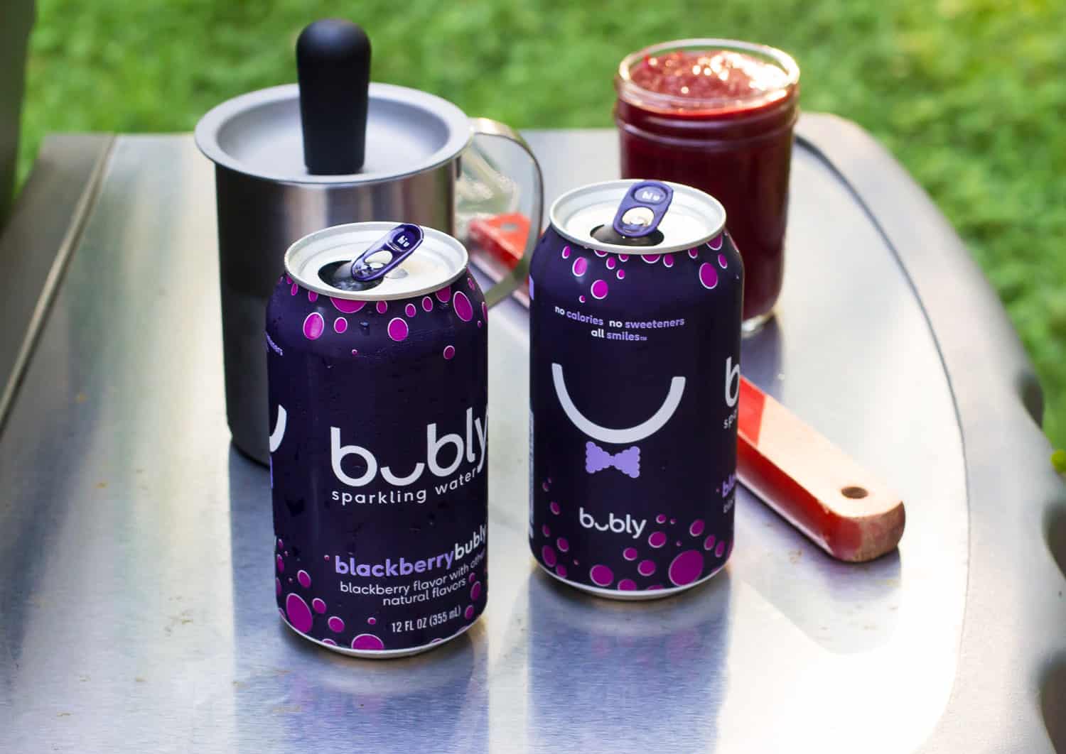 Sparkling water cans, a jar of BBQ sauce, and a spatula on the side table of a grill.