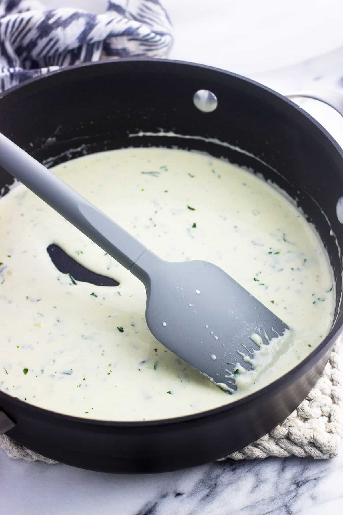 A skillet with tall sides containing the thickened cream sauce before adding the pasta, with a spatula in the middle of it