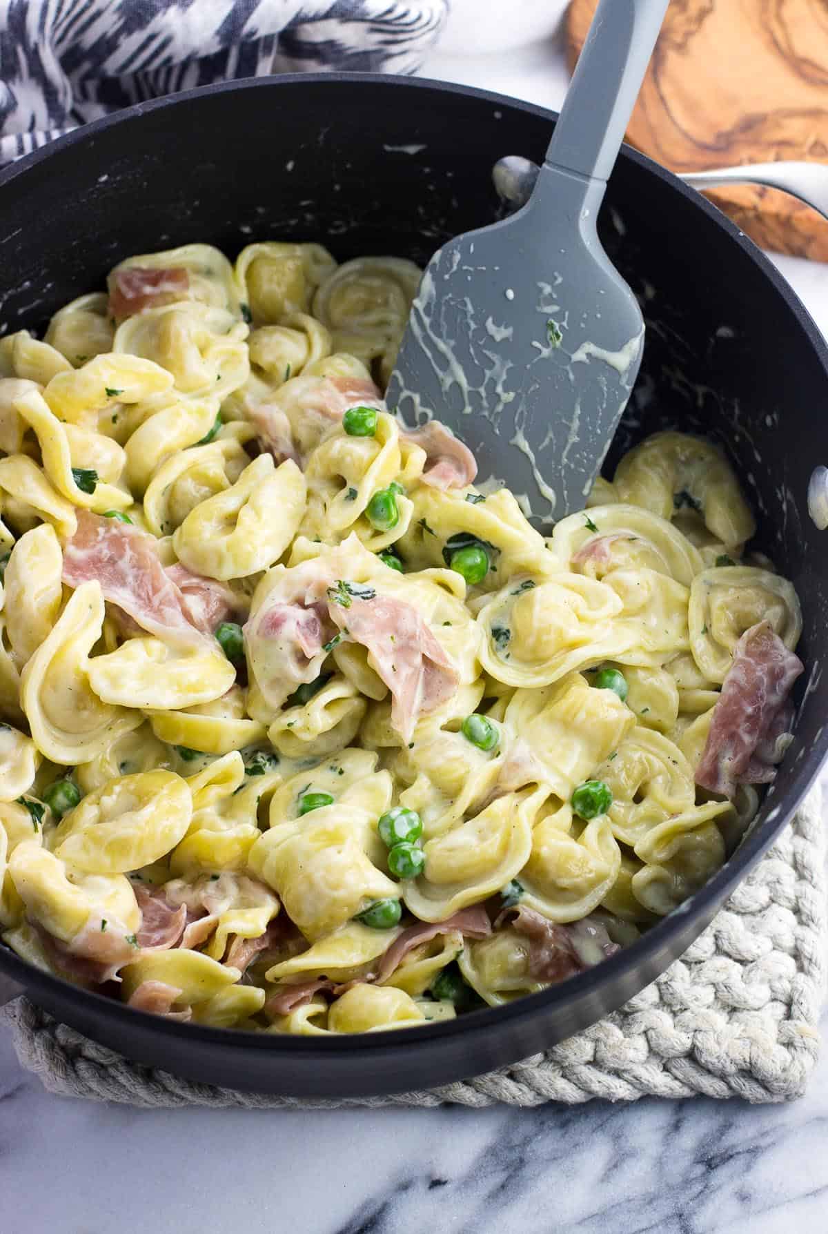 A pan filled with tortellini, prosciutto, and peas in a cream sauce with a spatula in it
