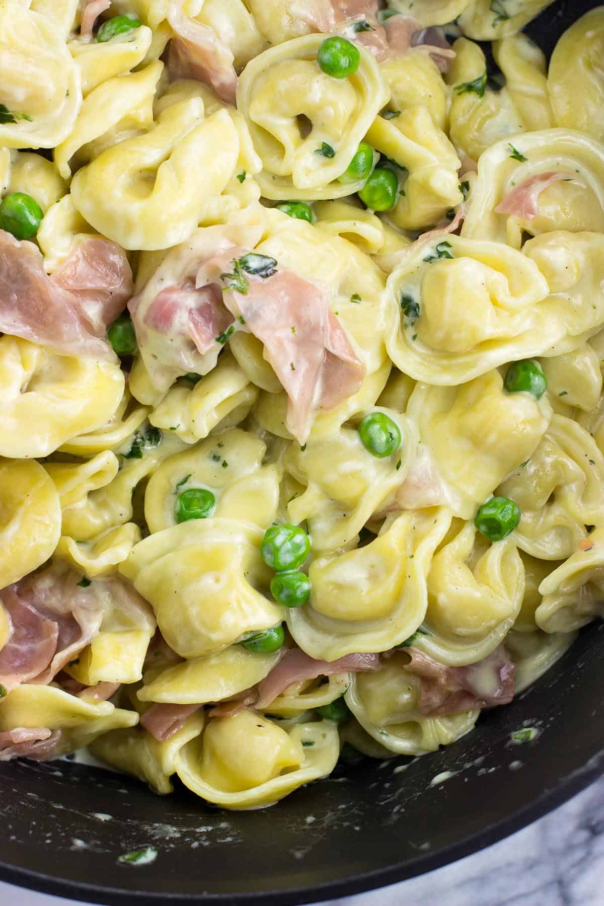 An overhead shot of the tortellini, prosciutto, and peas coated in the cream sauce in the skillet