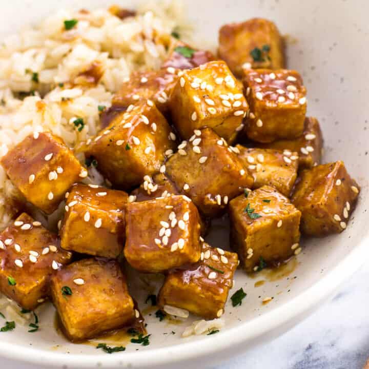 Cubes of tofu tossed in glaze and topped with sesame seeds in a shallow dinner bowl with brown rice
