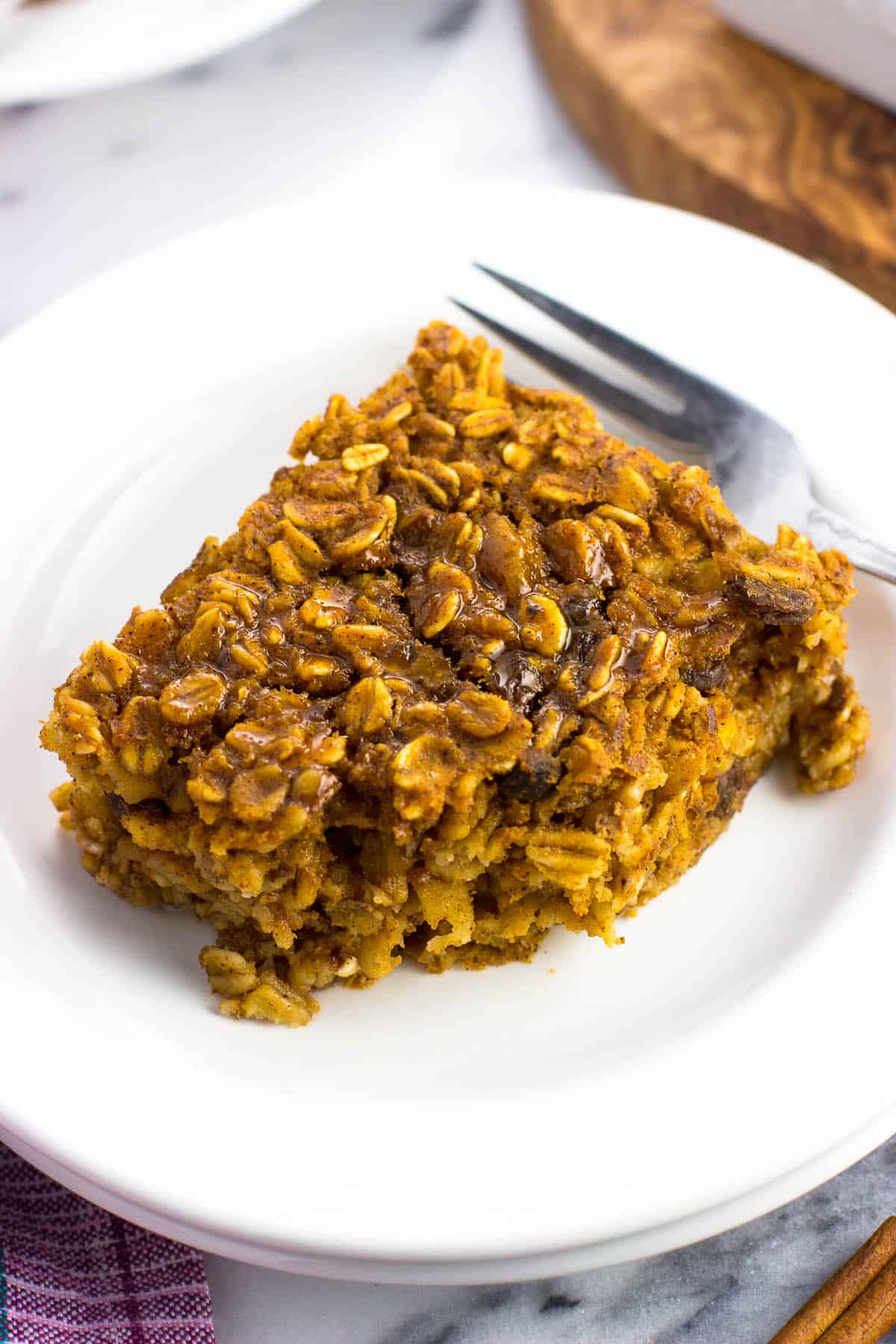 A close-up picture of a square of baked oatmeal with a bite taken out of it, drizzled in syrup, on a small plate with a fork