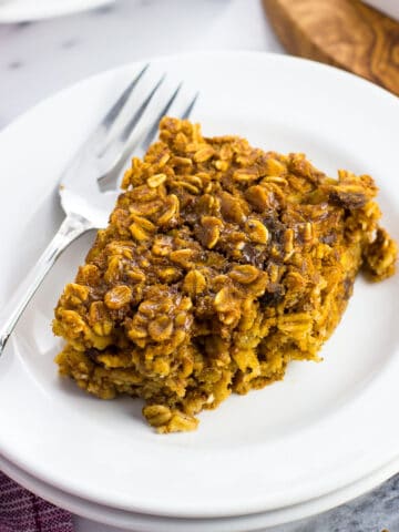 A square oatmeal bar on a small dessert plate with a fork with a bite taken out of it. Another serving is on a plate in the background