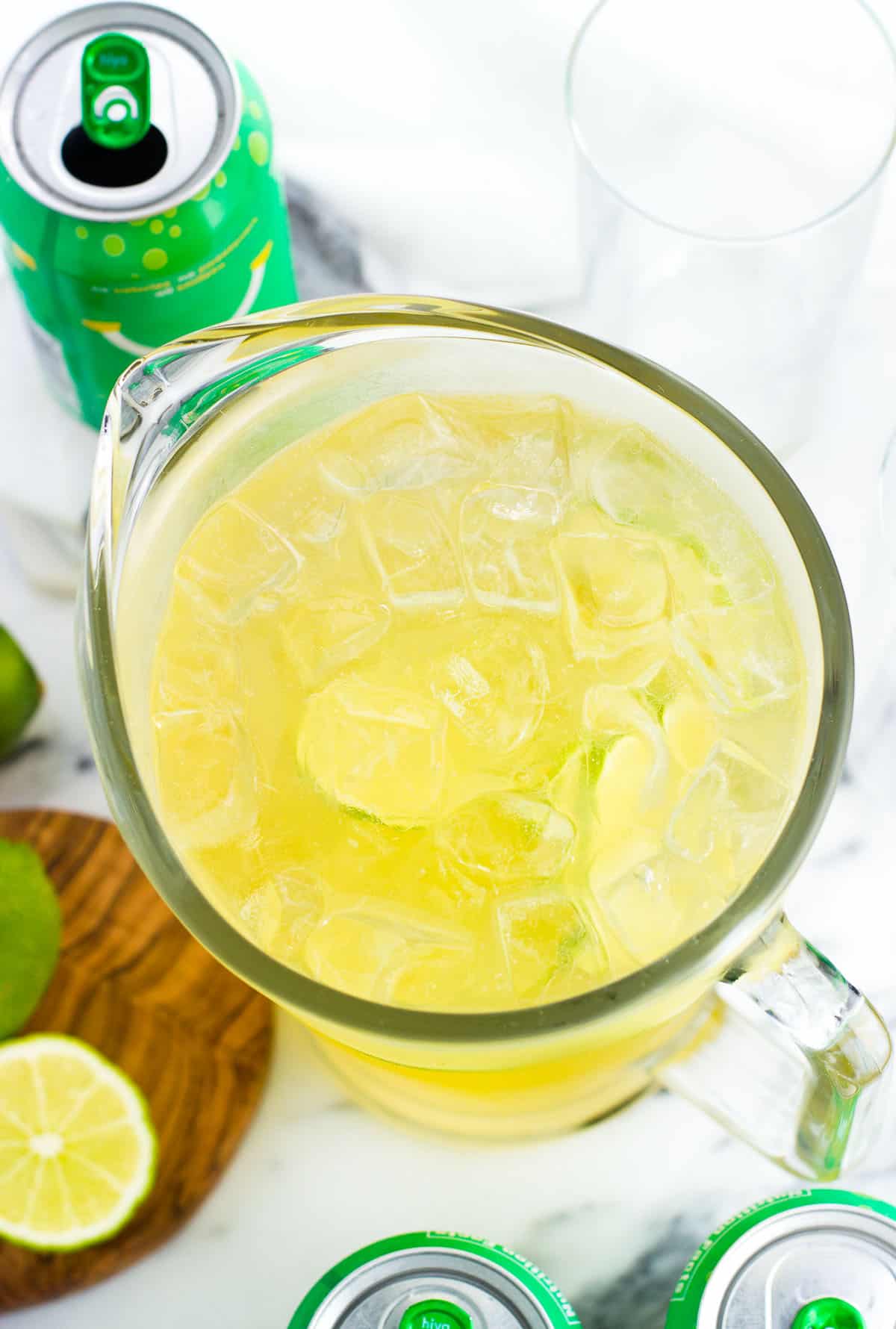 A glass mocktail pitcher filled with ice and next to cans of bubly and cut limes.