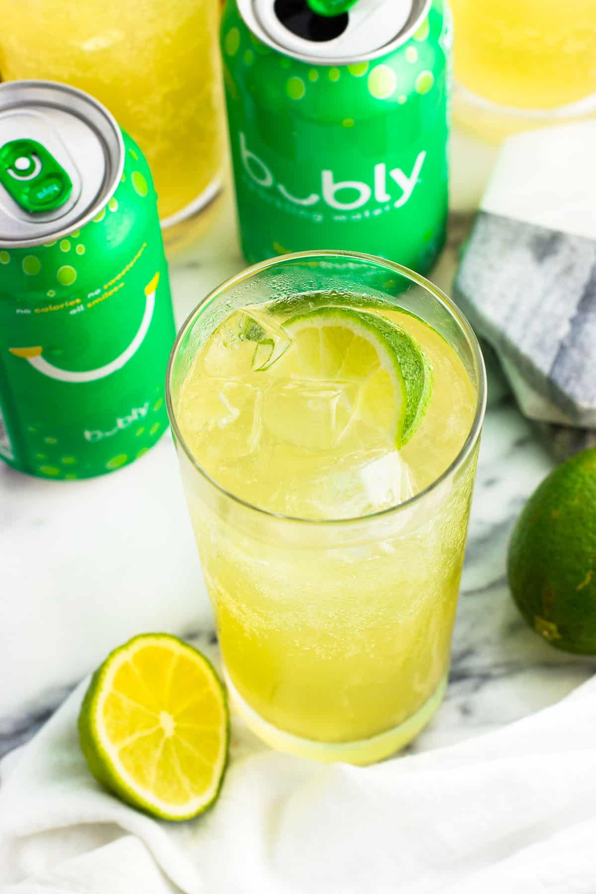 A close-up of a mocktail in a tall glass filled with ice and a lime wedge, with lime sparkling water cans surrounding