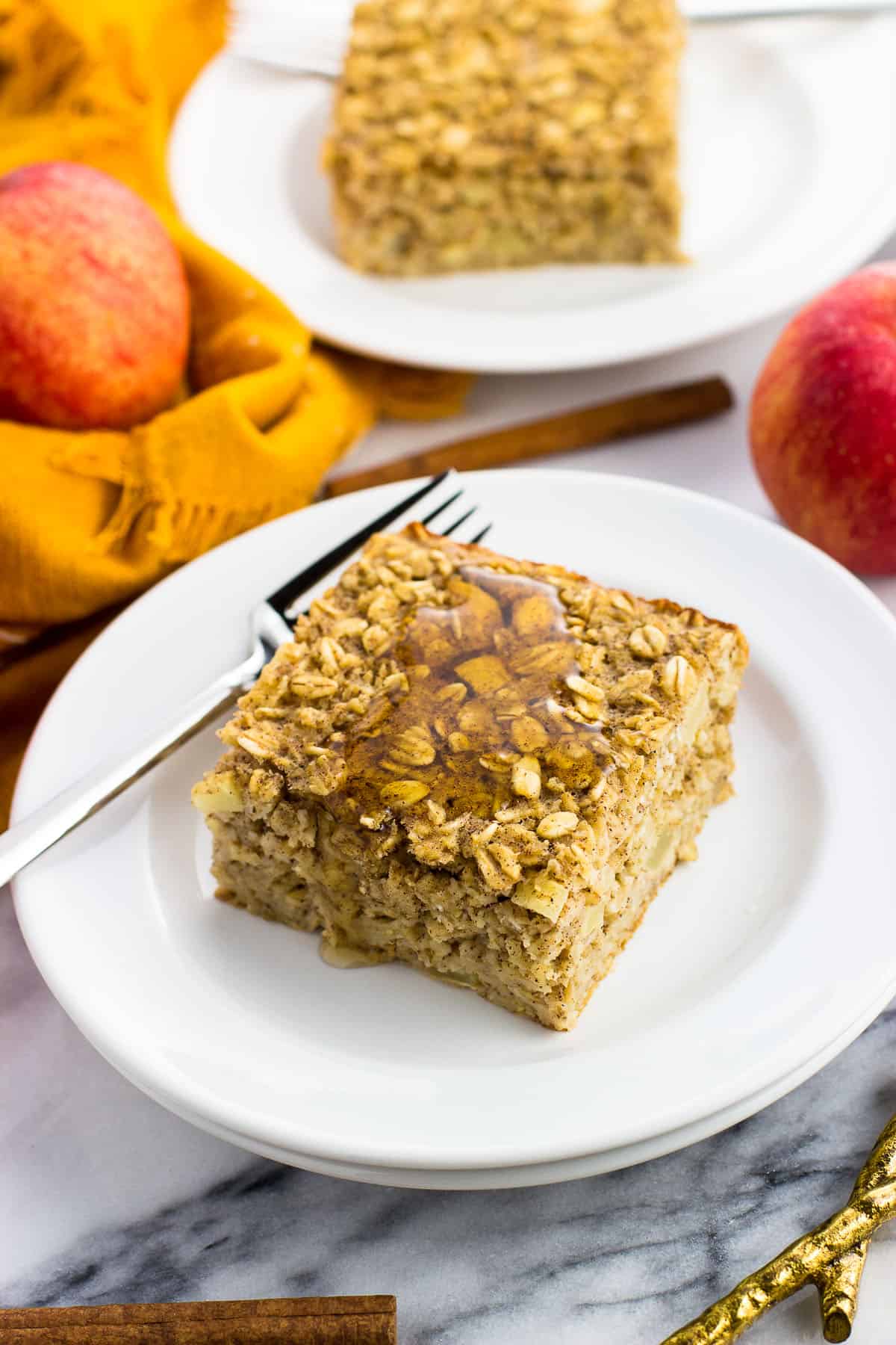 A piece of apple baked oatmeal on a plate with a fork next to a linen napkin, apples, and a cinnamon stick.