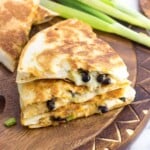 A stack of three quesadilla wedges on a wooden board with a bunch of green onions in the background