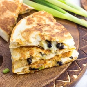 A stack of three quesadilla wedges on a wooden board with a bunch of green onions in the background