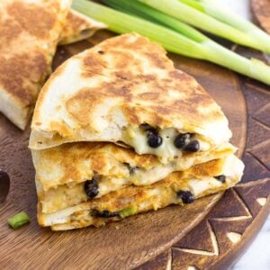 A stack of three quesadilla wedges on a wooden board with a bunch of green onions in the background