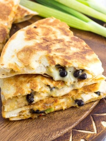 A stack of three quesadilla wedges on a wooden board with a bunch of green onions in the background