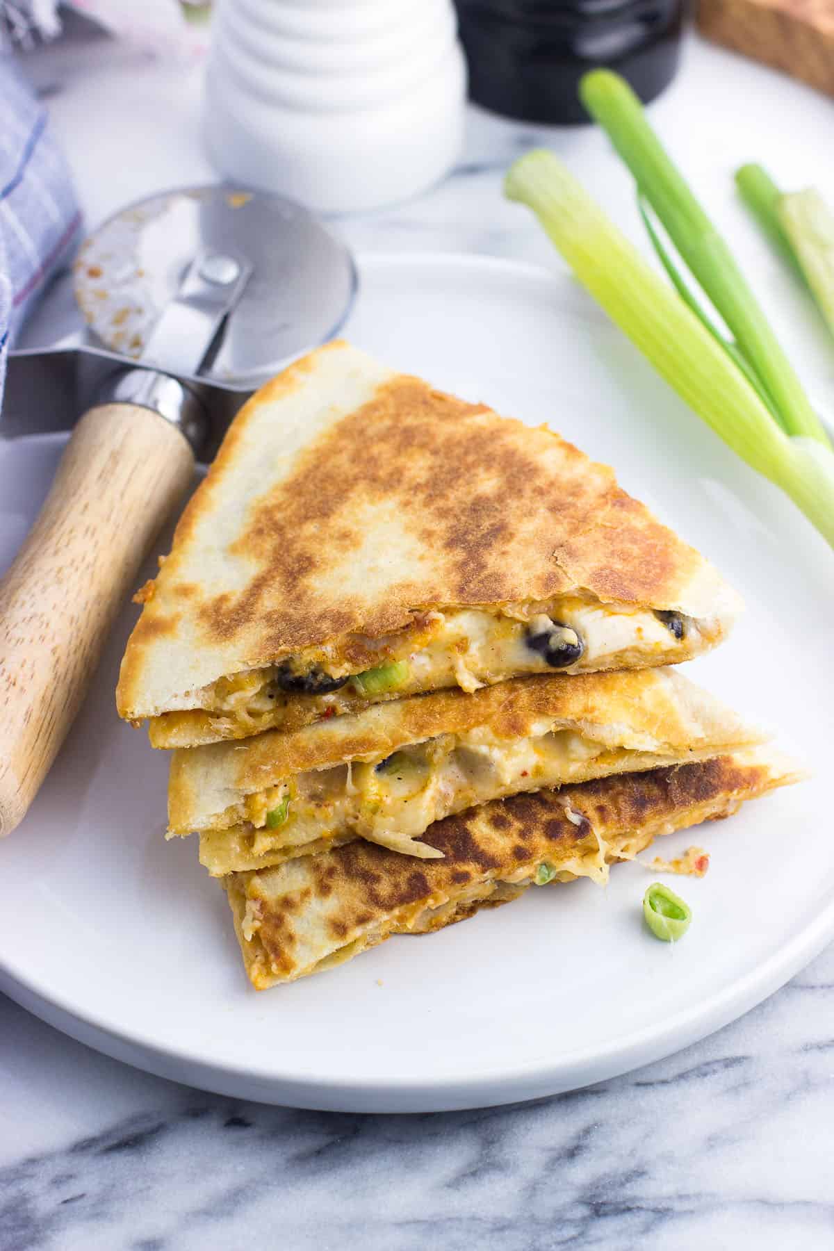 Three quesadilla wedges stacked on top of one another on an appetizer plate next to a pizza cutter and a bunch of green onions