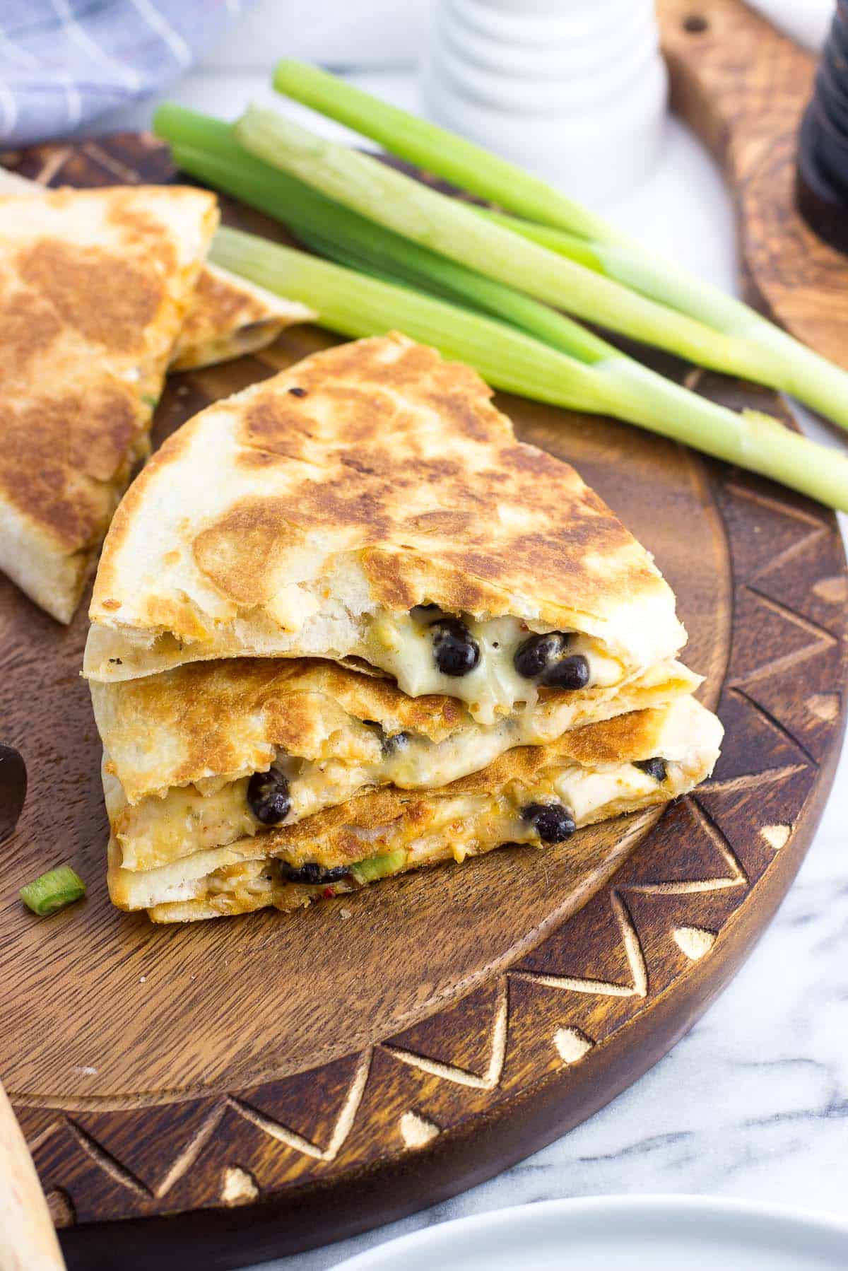 A stack of three quesadilla wedges on a wooden board with a bunch of green onions in the background