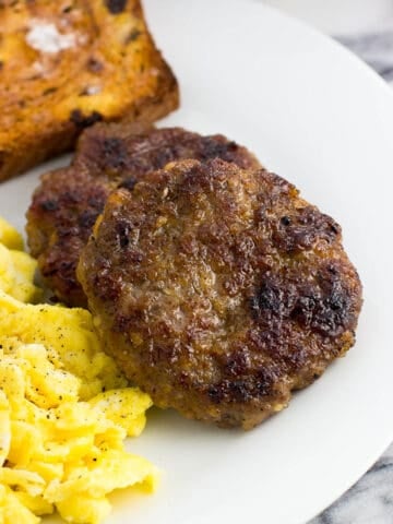 Two sausage patties on a plate next to scrambled eggs and a piece of cinnamon raisin toast