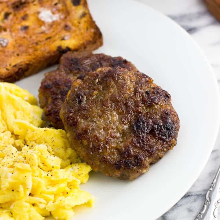 Two sausage patties on a plate next to scrambled eggs and a piece of cinnamon raisin toast
