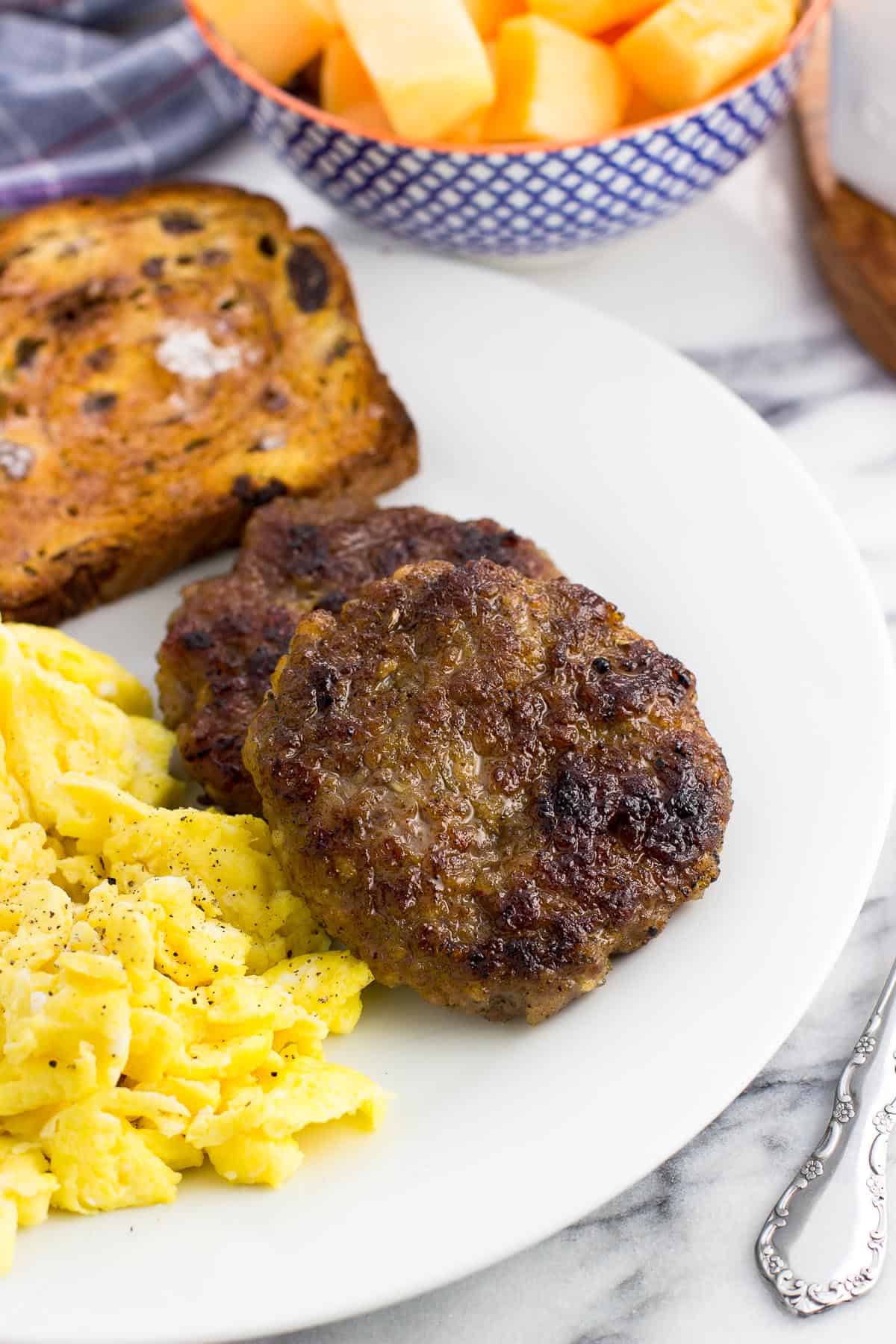 Two sausage patties on a plate next to scrambled eggs and a piece of cinnamon raisin toast with a bowl of cut up cantaloupe in the background