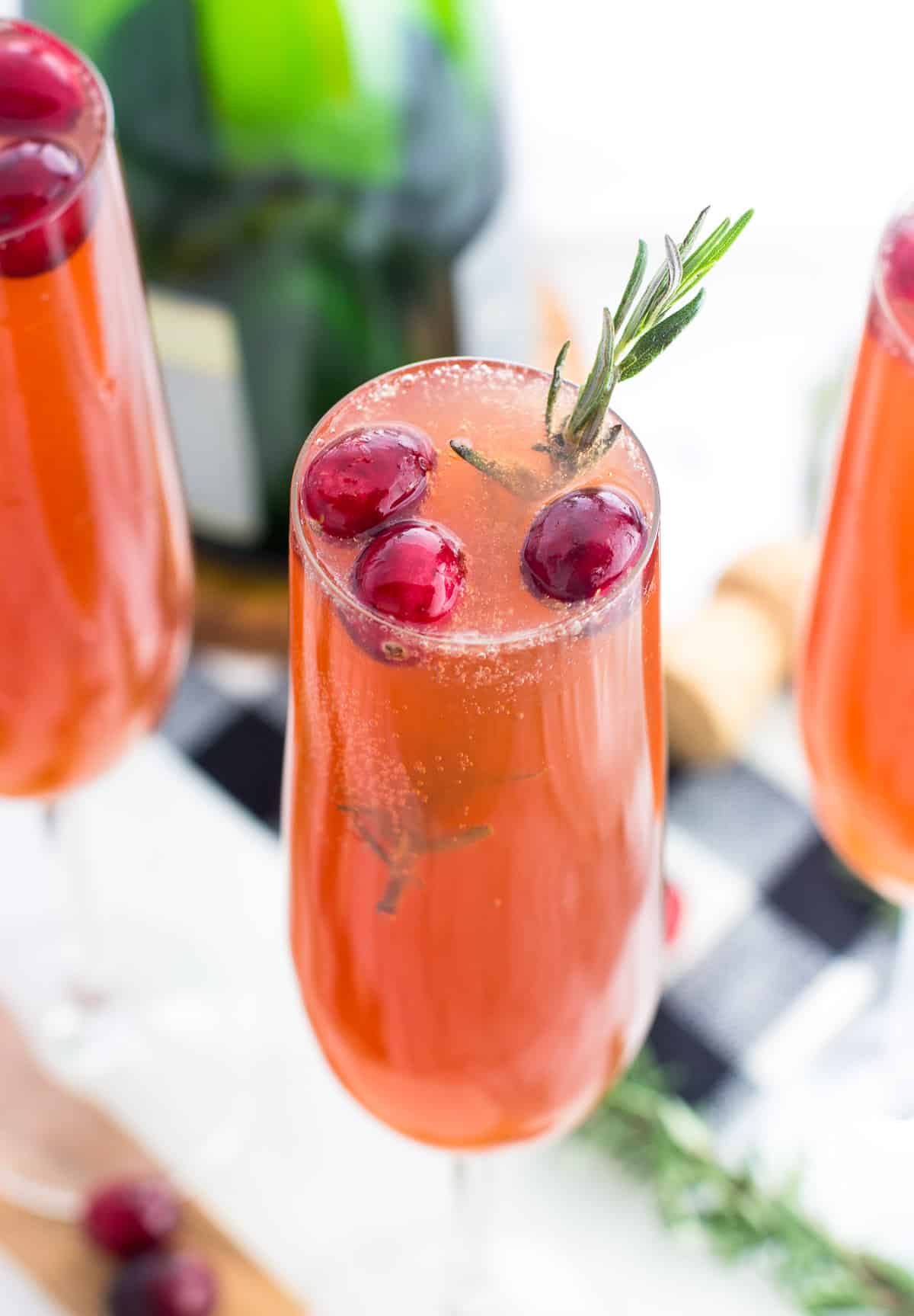 A close-up of a cranberry mimosa with three fresh cranberries and a sprig of rosemary for garnish.