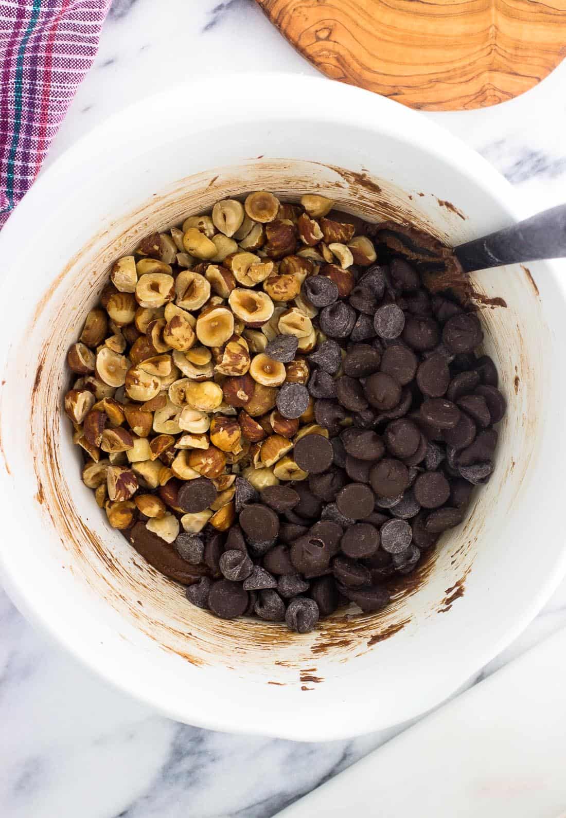 A bowl of brownie batter topped with chopped hazelnuts and chocolate chips ready to be mixed in