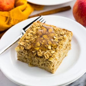 A piece of apple baked oatmeal on a plate with a fork next to a linen napkin, apples, and a cinnamon stick.