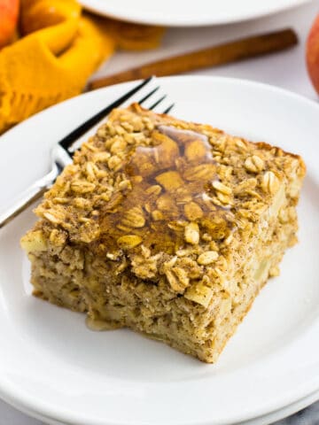 A piece of apple baked oatmeal on a plate with a fork next to a linen napkin, apples, and a cinnamon stick.