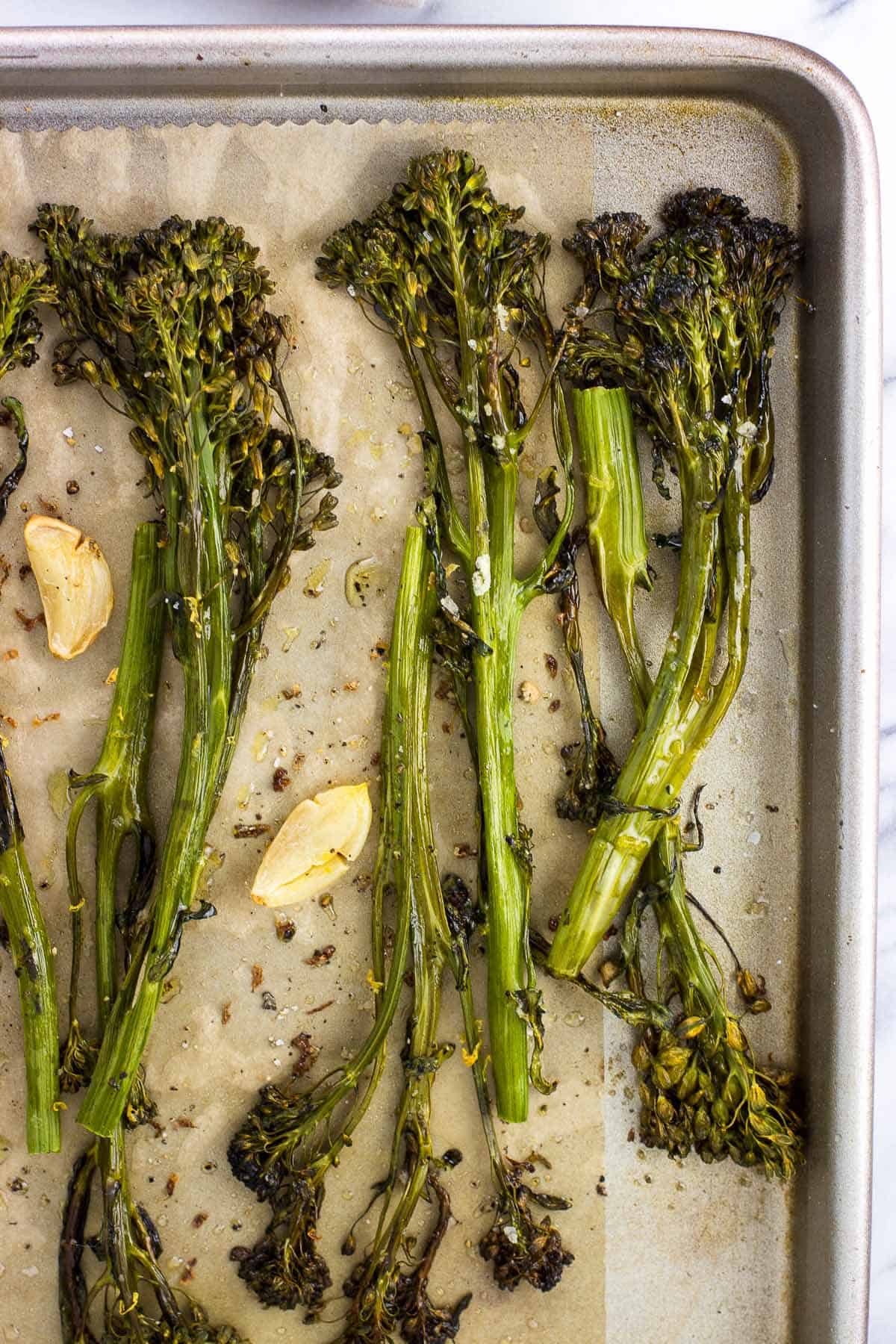 An overhead view of roasted broccolini spears and garlic cloves on a parchment-lined baking sheet.