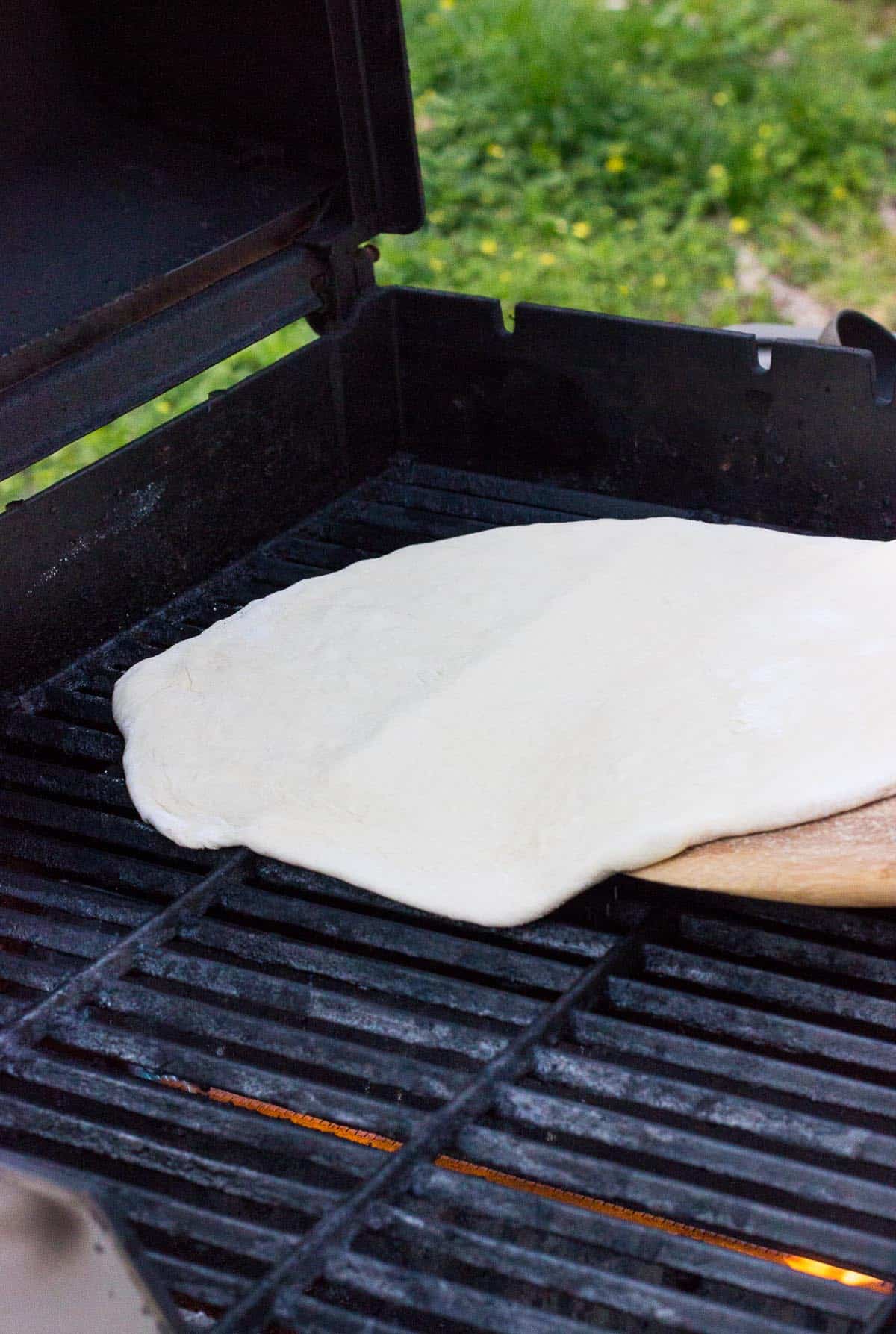 Stretched pizza dough being place on the grill by a pizza peel.