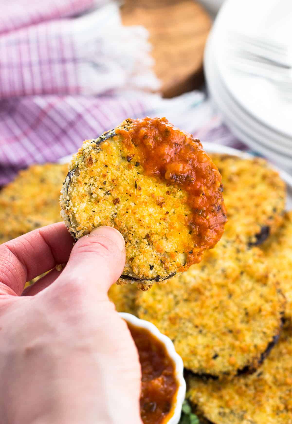 A hand holding up an eggplant round dipped in marinara sauce.