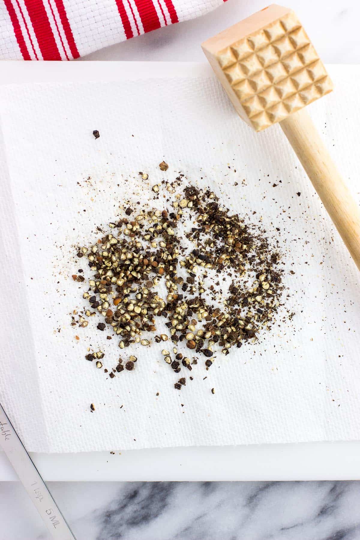 Smashed peppercorns on a paper towel next to a meat tenderizer.