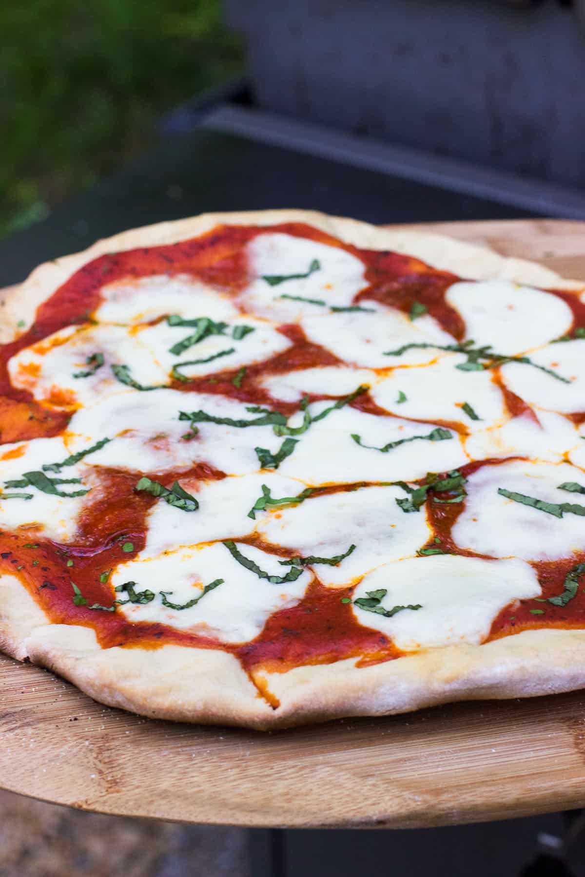 Grilled pizza on a wooden pizza peel on the side table of a grill.