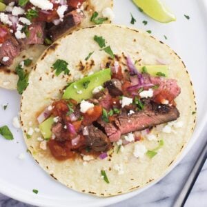 Carne asada tacos on a plate with avocado, salsa, and more toppings.