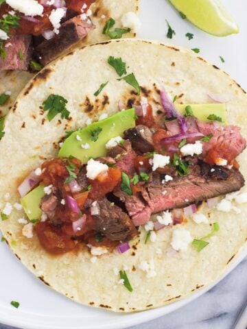 Carne asada tacos on a plate with avocado, salsa, and more toppings.
