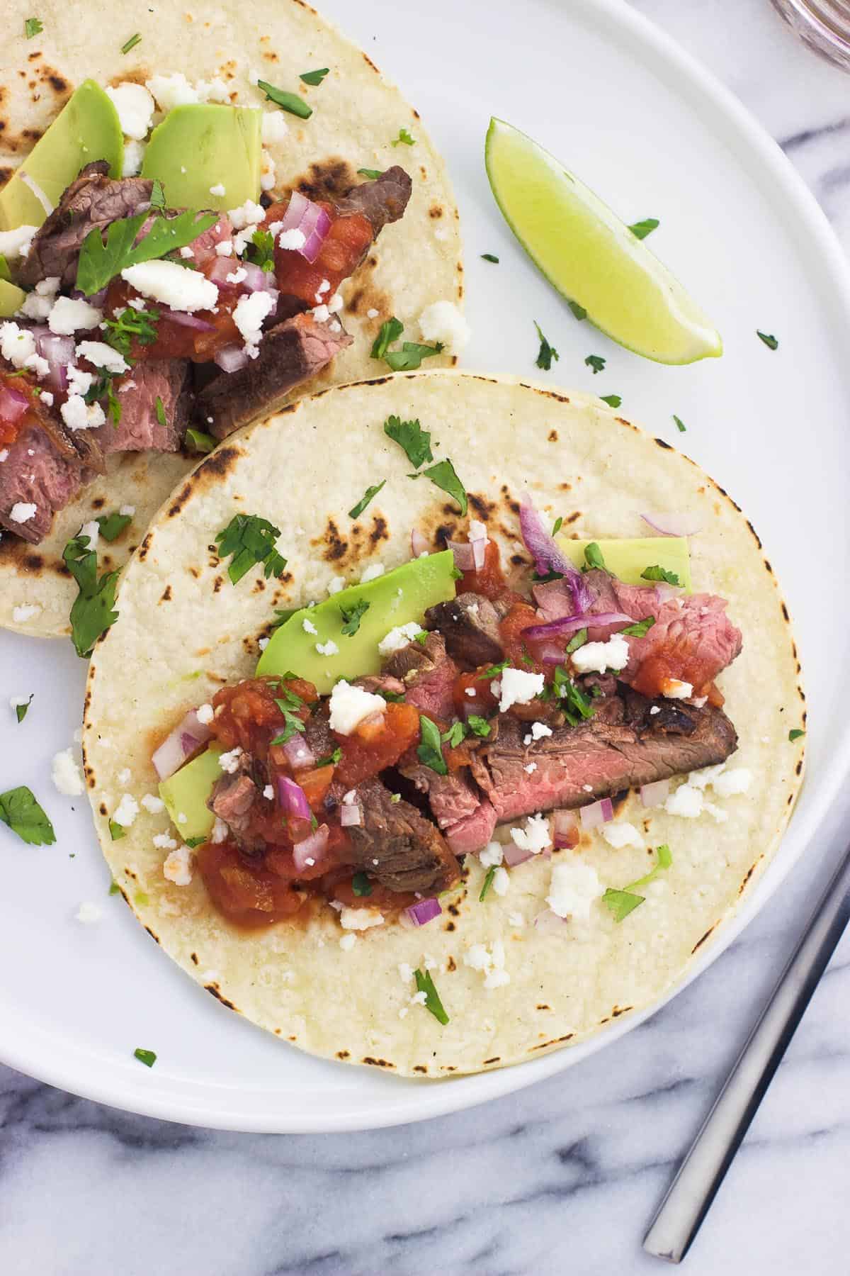 Two grilled carne asada tacos on a plate, including cotija cheese, salsa, avocado slices, and red onion.