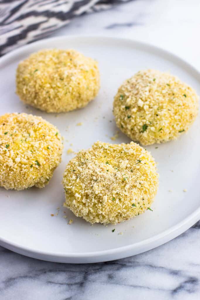 Four breadcrumb-coated risotto cakes on a plate before cooking.