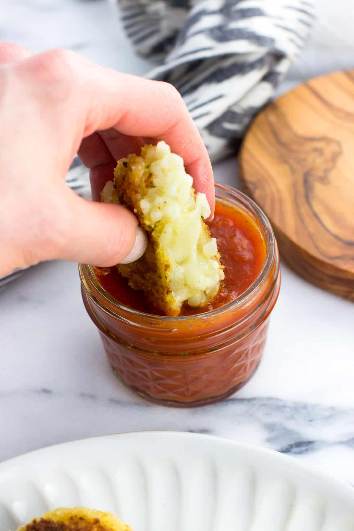 A hand dunking half of a risotto cake into a bowl of marinara sauce.