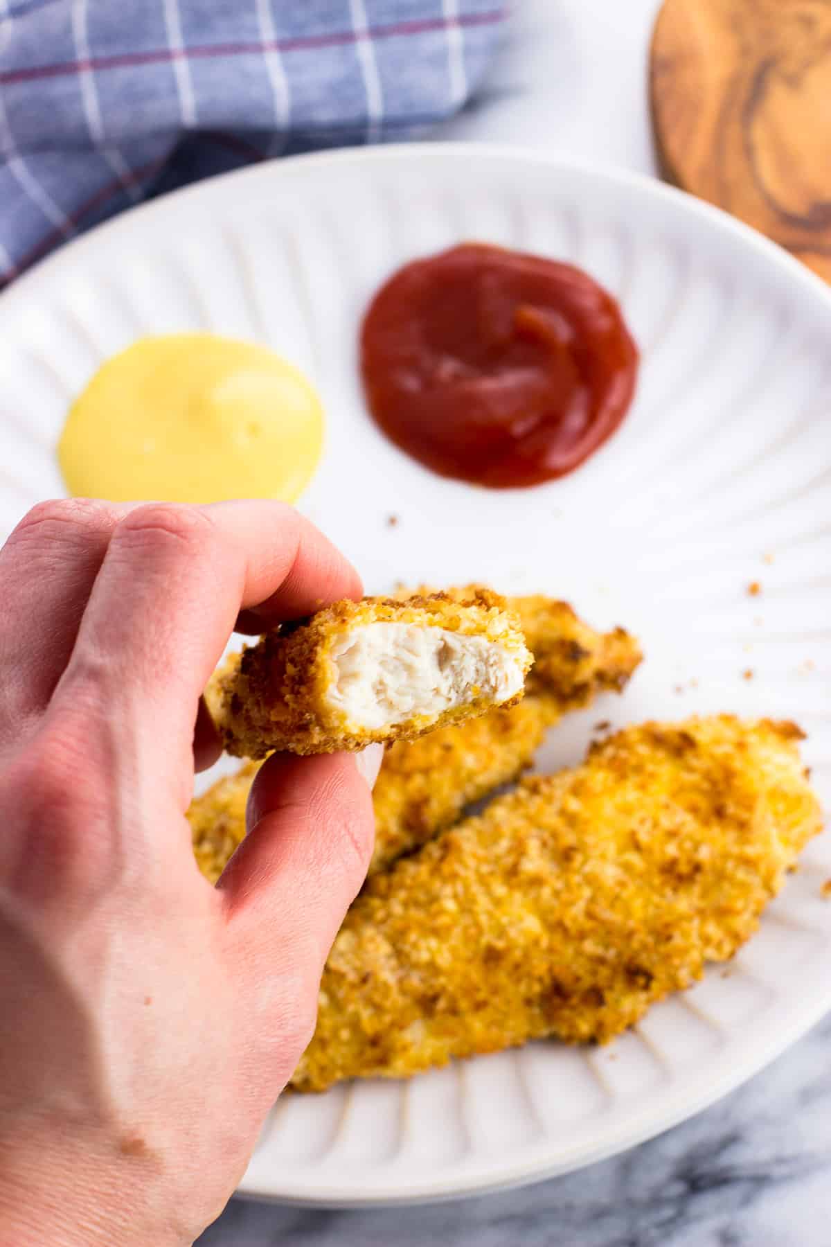 A hand holding a chicken tender with a bite taken out of it.