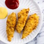 Three chicken tenders on a plate with ketchup and honey mustard.