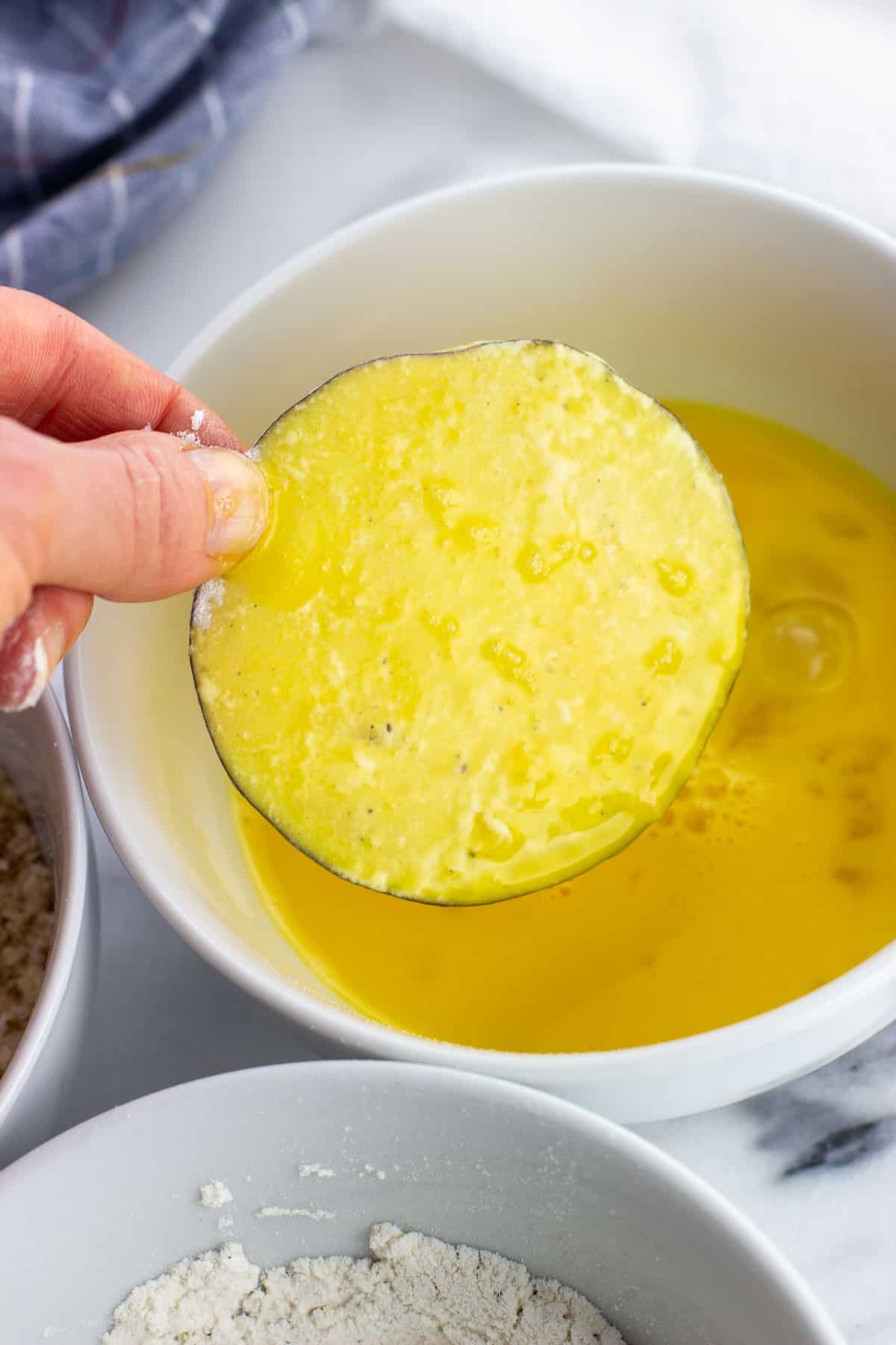 A hand holding an eggplant round dipped in egg above the bowl.