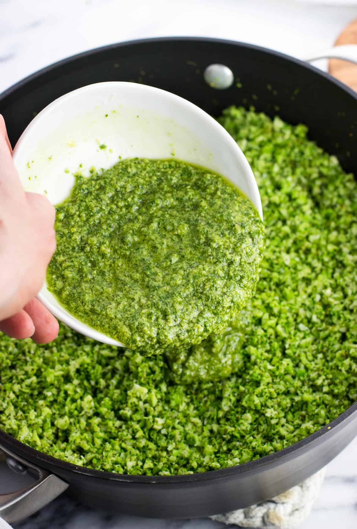 A hand pouring a bowl of pesto into the broccoli rice pan.