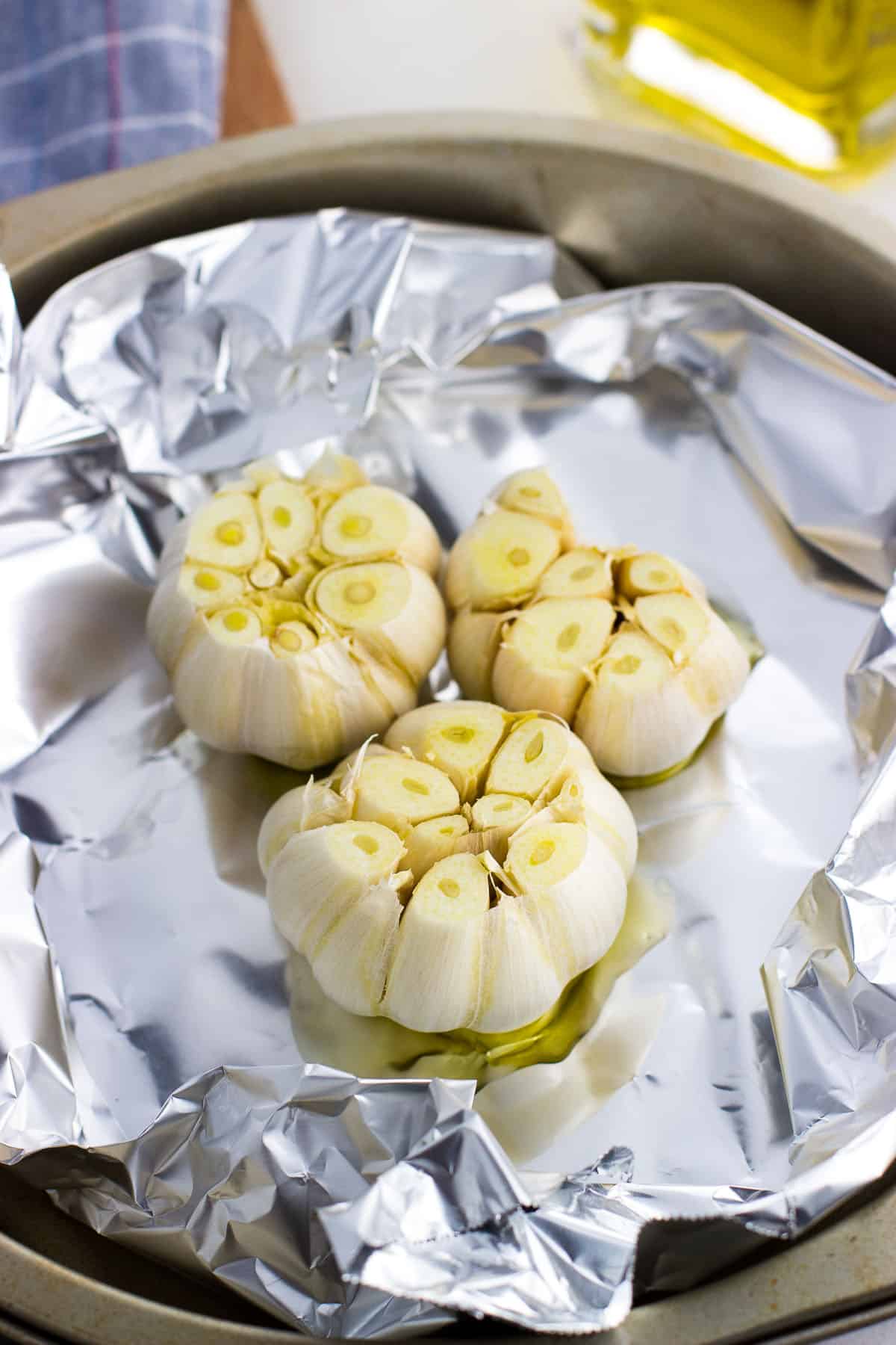 Three sliced and oil-drizzled garlic heads on foil before roasting.