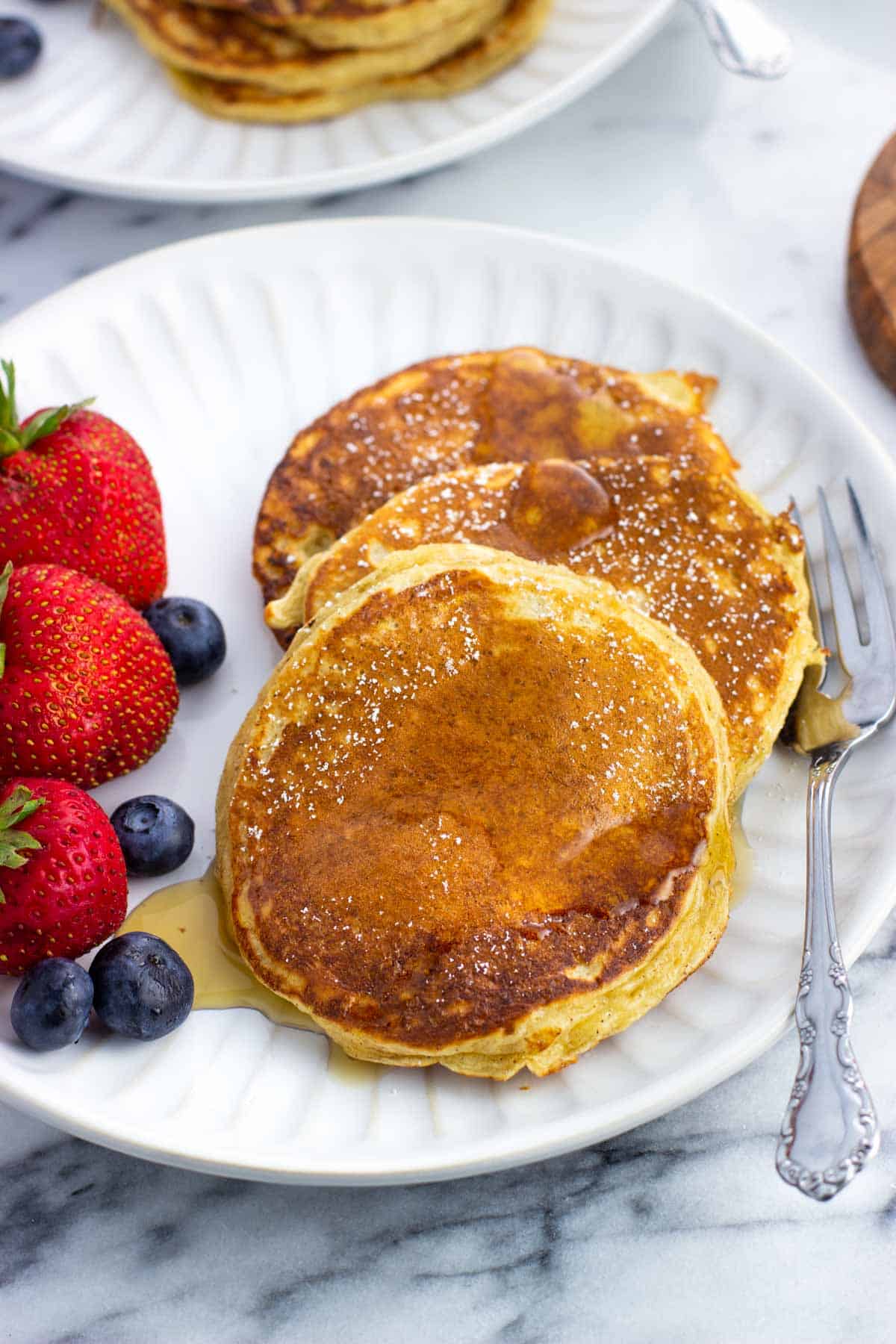 Three cottage cheese pancakes and berries on a plate drizzled with maple syrup.