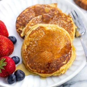 Three cottage cheese pancakes and berries on a plate drizzled with maple syrup.