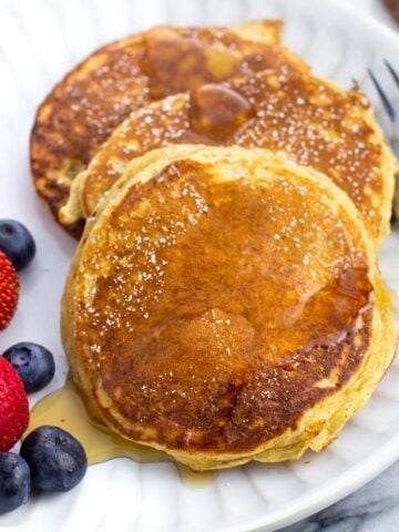 Three cottage cheese pancakes and berries on a plate drizzled with maple syrup.