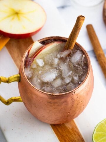 An apple cider mule garnished with apple slices and a cinnamon stick.