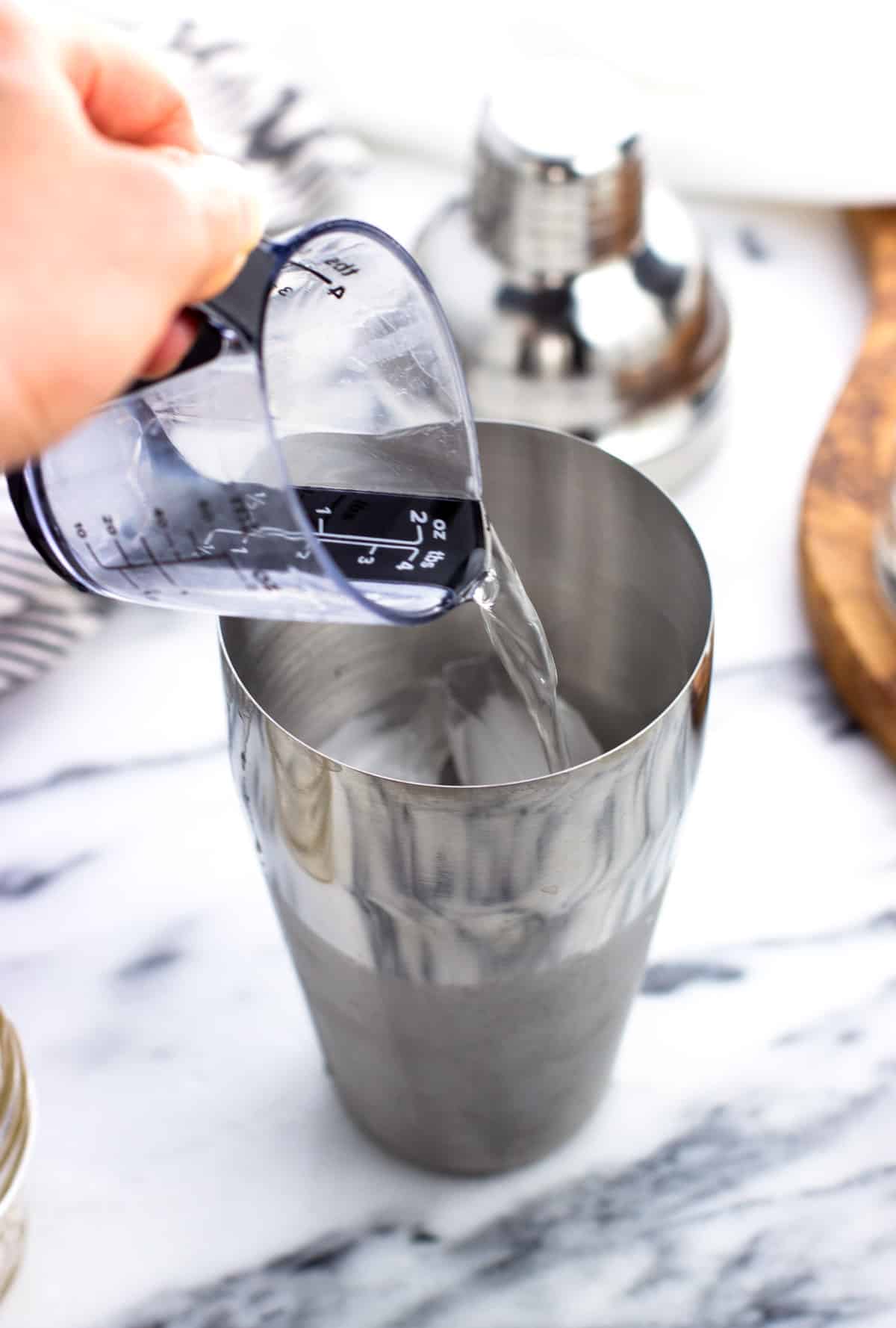 A hand pouring a small measuring cup of tequila into a cocktail shaker.