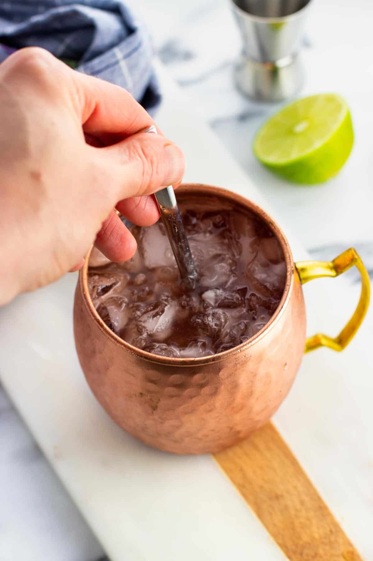 A hand stirring a cranberry Moscow mule with a spoon.