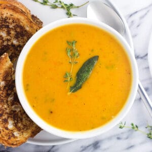 A bowl of butternut squash soup on a plate next to a grilled cheese.