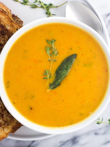 A bowl of butternut squash soup on a plate next to a grilled cheese.