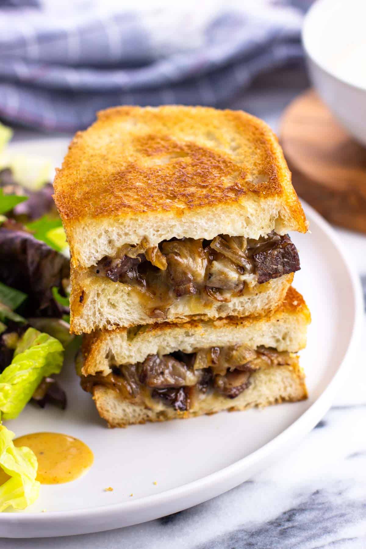 Two halves of a leftover steak sandwich stacked on top of one another on a plate.