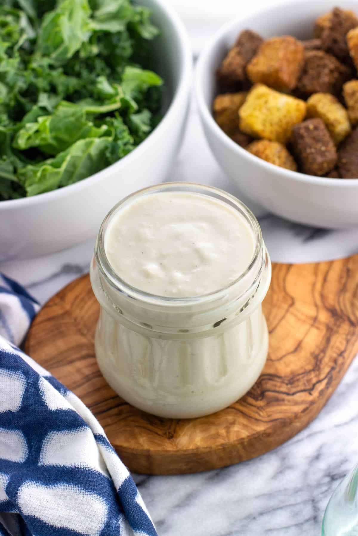 A glass jar of caesar dressing with salad ingredients in the background.