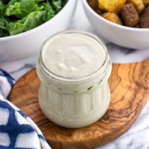A glass jar of Greek yogurt caesar dressing with salad ingredients in the background.