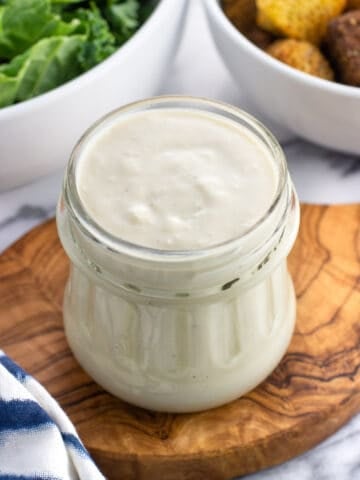 A glass jar of Greek yogurt caesar dressing with salad ingredients in the background.